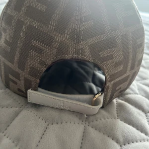 Beige Fendi keps med FF-mönster - Snygg keps från Fendi i beige och bruna toner med det ikoniska FF-logomönstret över hela kepsen. Justerbar rem bak i vitt läder med gulddetalj. Klassisk böjd skärm och vit knapp på toppen. Perfekt för dig som vill sticka ut med lyxig streetstyle.