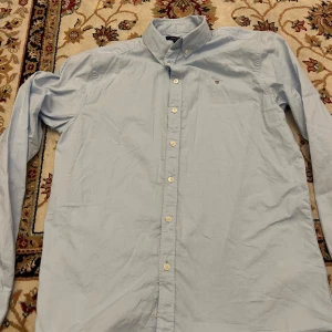 Ljusblå skjorta från Gant - Klassisk ljusblå skjorta från Gant med button-down krage och vita knappar. Broderad Gant-logga på bröstet. Skjortan är långärmad och tillverkad i mjuk bomull, perfekt för en clean och stilren look.