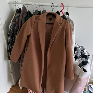 Brun kappa från ZARA med klassisk krage - Säljer en snygg brun kappa från ZARA med klassisk krage och rak passform. Jackan har långa ärmar och stilren design utan synliga knappar eller dragkedja. Perfekt för dig som vill ha en tidlös och trendig look.