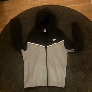 Nike tech hoodie i svart och grå med dragkedja - Nike tech hoodie i svart och ljusgrått med vit logga på bröstet. Tröjan har hel dragkedja, huva och långa ärmar. Snygg kontrast mellan svart upptill och grått nedtill, samt vit detalj längs bröstet. Perfekt för chill eller träning.