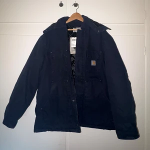 Mörkblå Carhartt jacka med huva L - Säljer en mörkblå Carhartt Ambel Coat i storlek L, endast provad. Jackan har huva, flera fickor och quiltat foder på insidan. Tillverkad i slitstark bomull och stängs med dragkedja. Perfekt för kalla dagar och har en clean streetwear-look.