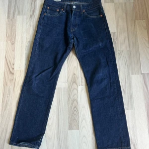 Levi's 501 mörkblå jeans W27 L28 - Klassiska Levi's 501 jeans i mörkblå denim med raka ben och femficksmodell. Byxorna har knappar i gylfen, kontrastsömmar och den ikoniska läderpatchen bak i midjan. Tillverkade i robust bomullsjeans för en tidlös look.