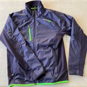 Blå vindjacka Polo Sport Ralph Lauren M - Sportig marinblå vindjacka från Polo Sport Ralph Lauren med gröna detaljer och dragkedjor. Jackan har hög krage, bröstficka med grön dragkedja och grönt kantband nertill. Tillverkad i polyester och passar perfekt för aktiva dagar.