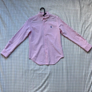 Rosa skjorta från Polo Ralph Lauren - Snygg rosa skjorta från Polo Ralph Lauren med klassisk krage och blå broderad logga på bröstet. Skjortan har lång ärm, knappar framtill och är gjord i mjuk bomull. Perfekt för dig som gillar stilrena och fräscha plagg med ikonisk design.