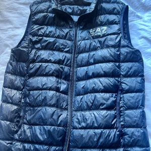 Marinblå dunväst från Emporio Armani EA7 - Snygg marinblå dunväst från Emporio Armani EA7 med quiltad design och dragkedja framtill. Västen har hög krage och logga tryckt på bröstet. Nypris 2000kr. Använda 3-4 gånger 
