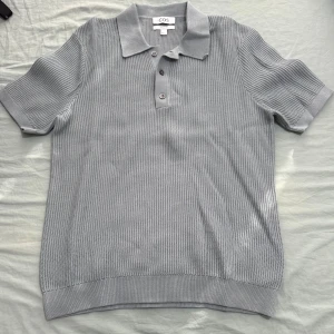 Grå stickad pikétröja från COS - Säljer en stilren grå stickad pikétröja från COS i storlek S. Tröjan har klassisk krage, tre knappar framtill och korta ärmar. Materialet är mjukt och strukturerat vilket ger en modern känsla. Perfekt för dig som gillar minimalistisk stil.