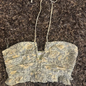 Blommig bandeau topp med volang - Supersöt bandeau topp med blommigt mönster i gröna och gula toner. Toppen har volangkant nertill, smockad struktur och knytband i nacken. Perfekt för varma dagar när du vill ha en romantisk och trendig look.