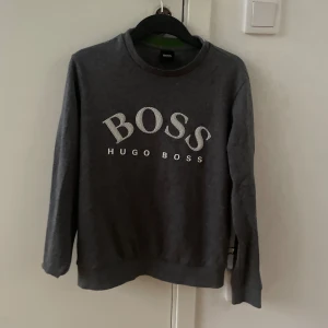 Mörkgrå sweatshirt från Hugo Boss - Snygg mörkgrå sweatshirt från Hugo Boss med stort vitt BOSS-tryck på bröstet. Klassisk rund halsringning och långa ärmar. Perfekt för en stilren och avslappnad look. Storlek Xs