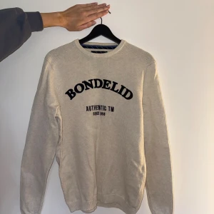 Beige stickad tröja från Bondelid - Säljer en beige stickad tröja från Bondelid med svart text på bröstet. Tröjan har rund halsringning, ribbade muddar och är långärmad. Perfekt för lager-på-lager och chill stil. Modellen heter Sergio Crew Neck.