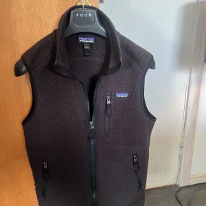 Svart fleeceväst från Patagonia - Svart fleeceväst från Patagonia i storlek S. Västen har hög krage, tre praktiska dragkedjefickor och hel dragkedja framtill. Perfekt för lager-på-lager och outdoor-aktiviteter. Mjuk och varm med klassisk Patagonia-logga på bröstet. Priset är inte hugget i sten.