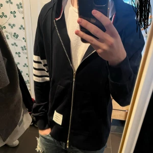  hoodie från Thom Browne  - Inte fast pris! Blå hoodie från Thom Browne med guldfärgad dragkedja, huva och snörning. Klassiska vita ränder på ena ärmen och fickor framtill. Tillverkad i bomull och har en snygg label på magen. Perfekt för dig som gillar stilrena streetwear-vibbar.