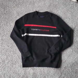 Svart Tommy Hilfiger sweatshirt XL - Svart sweatshirt från Tommy Hilfiger i storlek XL. Tröjan har broderad logga och text framtill, samt röda och vita ränder över bröstet. Klassisk rund halsringning och långa ärmar. Perfekt för en stilren och avslappnad look.