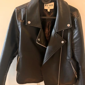 Svart skinnjacka från NA-KD, stl 34 - Snygg svart skinnjacka från NA-KD med klassisk bikerstil. Jackan har silvriga dragkedjor, bred krage och coola detaljer med tryckknappar. Storlek 34 men har lite större passform så passar även för dem som bär stk 36-38.