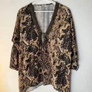Frontrow - Elegant tunika från Frontrow med ett unikt ormskinnsmönster i svart, beige och rosa. V-ringningen är vackert dekorerad med guldfärgade detaljer och har en justerbar knytning framtill. Denna tunika har en avslappnad passform och storlek M