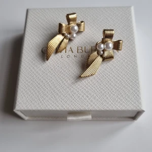 Guldiga örhängen med pärlor - Säljer ett par snygga örhängen i guldtonad metall med rosettformad design och vita pärlor som detalj. De har en cool struktur och fästs med klassisk stift. Perfekt för dig som gillar statement-smycken med lyxig vibe. Lådan ingår ej.