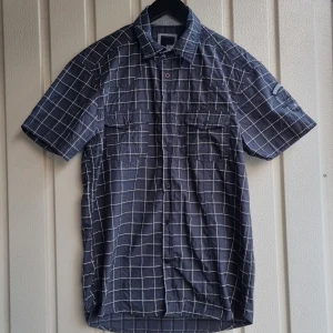 Rutig blå skjorta från Jack & Jones - Kortärmad skjorta från Jack & Jones i storlek XL. Skjortan är mörkblå med vitt rutmönster. Förutom en tråd som har gått upp och en annan defekt, så har tröjan knappt används.