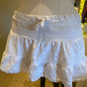 Söt volangkjol med spets - Supersöt vit kortkjol med volang och spetsdetaljer. Kjolen har knyt i midjan och ett luftigt, stickat material som känns perfekt för varmare dagar. Den feminina looken förstärks av de små hålmönstren och den vågiga nederkanten. Mindre i storleken💞