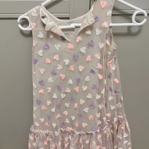 Beige klänning med glitterhjärtan - Supersöt beige ärmlös klänning med volangkant nertill och massor av glittriga hjärtan i lila, vitt och rosa. Klänningen har en lätt och luftig känsla, perfekt för dig som gillar gulliga detaljer och pastellfärger.