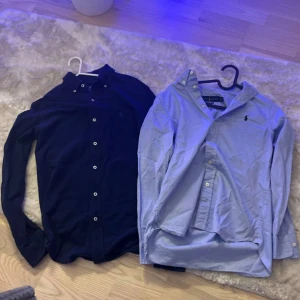 Två skjortor från Ralph Lauren - Två klassiska skjortor från Ralph Lauren, en i mörkblått och en i ljusblått. Båda har button-down krage, långa ärmar och knappar framtill. Ikoniska broderade loggan på bröstet. Perfekta för en clean och stilren look.