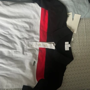 Lacoste piké med röd och svart detalj - Snygg pikétröja från Lacoste i bomull med klassisk krage och knappar framtill. Skjortan har vita, svarta och röda blockfärger samt den ikoniska krokodilloggan på bröstet. Perfekt för en stilren och sportig look.