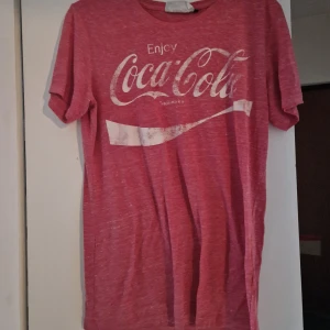 Röd Coca-Cola t-shirt i bomull - Röd t-shirt från Coca-Cola med klassiskt vitt tryck framtill. T-shirten är kortärmad och har en avslappnad passform. Materialet är mjuk bomull och färgen är melerad röd med vita detaljer. Perfekt för dig som gillar retrostil och ikoniska loggor.
