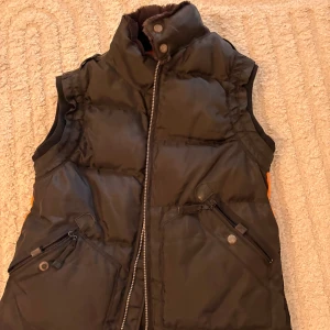 Svart dunväst från Parajumpers - Svart dunväst från Parajumpers Masterpiece Series Anthology med orange detaljer och broderad patch på axeln. Västen har hög krage med mjuk fuskpäls, dragkedja framtill och två snedställda fickor med dragkedja och knapp. Perfekt för kalla dagar.