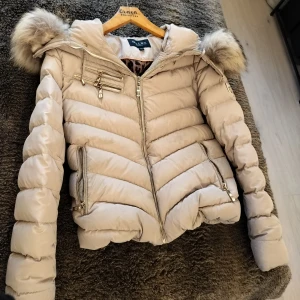 Beige Hollies jacka med guldiga detaljer  - Snygg beige pufferjacka från Hollies med tvättbjörns päls på huvan och guldfärgade dragkedjor. OBS! Lite fläckig här och där, behövs kemtvättas. Strl: 40 (Känns som 36/38 dock)