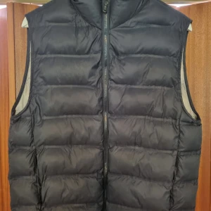 Svart dunväst från Zara - Säljer en svart, quiltad dunväst från Zara med dragkedja framtill. Västen har en hög krage och är fodrad i beige. Perfekt för lager-på-lager och enkel att matcha med olika outfits.