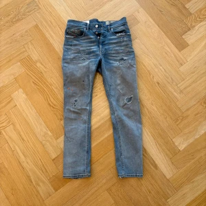 Blå slitna jeans från Diesel 30 32 - Säljer ett par blå jeans från Diesel med coola slitningar, hål och färgstänk i orange och svart. Jeansen har klassisk femficksdesign, raka ben och tydliga detaljer som ger en edgy vibe. Perfekta för dig som gillar streetstyle och vill sticka ut.