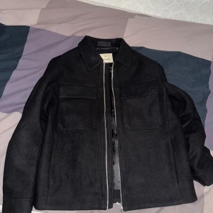 Overshirt från Jack & Jones - Stilren svart overshirt från Jack & Jones med klassisk krage, två stora bröstfickor och dragkedja framtill. Yttermaterialet är i ullblandning och insidan har ett lent, glansigt foder. Perfekt för lager-på-lager och enkel att matcha. Helt oanvänd och säljs för att den var för stor! Fraktas endast och köparen står för frakten!