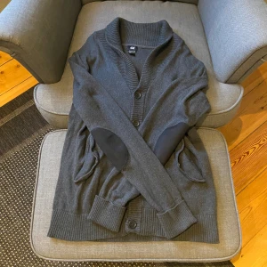 Mörkgrå kofta från H&M med knappar - Mysig mörkgrå kofta från H&M med bred ribbad krage, knäppning framtill och förstärkta armbågslappar. Koftan har två fickor och ribbade muddar vid ärmslut och nederkant. Perfekt för lager-på-lager under kyliga dagar.