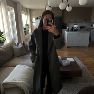 Grå/grön kappa med bälte från Zara - Säljer en lång, grå-grön kappa från Zara med bälte i midjan och breda slag. Kappan har en rak och loose passform, långa ärmar och är perfekt för höst och vinter. Enkel och stilren design som funkar till många outfits.