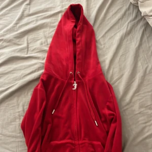 Röd velourhoodie från Juicy Couture - Juicy Couture hoodie i röd velour med dragkedja och huva. Klassisk J-dragkedja, snörning i huvan och broderad logga på bröstet. Perfekt för dig som vill ha en ikonisk och mjuk hoodie med 00-talsvibe.