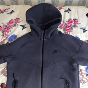 Mörkgrå Nike hoodiejacka med dragkedja - Nike Tech Fleece hoodiejacka i mörkgrått med hel dragkedja och två fickor med svarta dragkedjor. Jackan har en stor huva och diskret Nike-logga på bröstet. Perfekt för chill dagar eller när du vill ha en sportig look. Helt ny! Använd 1 gång bara.