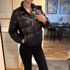 Svart dunjacka från Levi's, M - Snygg svart dunjacka från Levi's med glansig finish och klassisk pufferstil. Jackan har dragkedja framtill, hög krage och Levi's-logga på bröstet. Vattenavvisande material och värmande dunfyllning gör den perfekt för kyliga dagar nu när hösten kommit. Medföljer även en liten ”påse” där man kan stoppa in jackan i så den knappt tar någon plats alls! Därav att jackan kanske ser lite skrynklig ut, då jag precis tog ut jackan ur påsen! ☺️ Den är i finfint skick! Pris kan diskuteras vid snabb affär! 