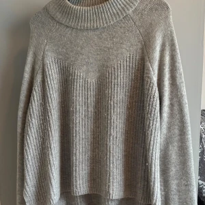 Grå stickad tröja från KappAhl - Mysig grå stickad tröja från KappAhl med bred ribbad krage och snyggt strukturerat mönster framtill. Perfekt för kyliga dagar och enkel att styla med jeans eller kjol.