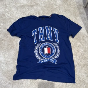 Blå t-shirt från Tommy Hilfiger - Blå t-shirt från Tommy Hilfiger med stort tryck framtill där det står THNY och en emblem med röd, vit och blå detalj. Tillverkad i ekologisk bomull och har klassisk passform med korta ärmar. Perfekt för dig som gillar sportig och stilren streetwear.