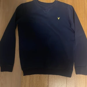 Mörkblå sweatshirt från Lyle & Scott - En stilren mörkblå sweatshirt från Lyle & Scott Junior med klassisk rund hals och gul broderad logga på bröstet. Tröjan har långa ärmar och ribbade muddar vid ärmslut och nederkant. Perfekt för en clean och enkel look.