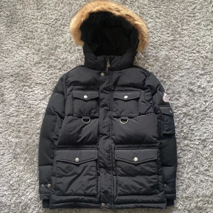 Svart dunjacka från Pyrenex med fuskpäls - Säljer en svart dunjacka från Pyrenex Heritage Collection med stor huva och fuskpäls. Jackan har fyra fickor framtill med lock och tryckknappar, samt två D-ringar. Klassisk vinterjacka med snyggt badge på ärmen och dragkedja framtill.