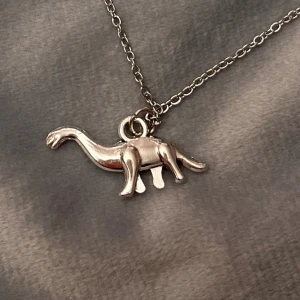 Halsband Dinosaurie Smycke Silver Imitation  - Så fint halsband med en berlock av en dinosaurie, en brontosaurus i detta fallet 🦕  Halsbandet är justerbart 40-45 cm och är i silverimitation. Berlocken är ca 2,5 x 1 cm. 