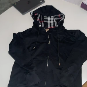 Svart Burberry hoodie XS med dragkedja - Svart hoodie från Burberry i storlek XS med klassiskt rutigt foder i huvan, broderad logga på bröstet och dragkedja framtill. Snygga metalldetaljer med Burberry-gravyr och justerbar snörning i huvan. Perfekt för dig som vill ha en stilren och lyxig look. Passar 176cm är xs men passar som s 
