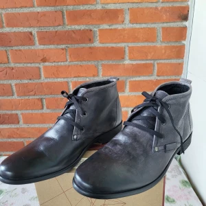 Svarta skinnkängor smoke grå från Legend - Snygga svarta kängor från Legend i äkta skinn med klassisk snörning och rund tå. Skorna har en stilren design med låg klack och svart sula, perfekta för dig som gillar en clean och tidlös look.