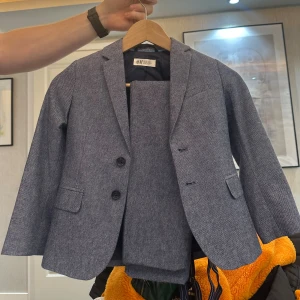 Blå kavajset från H&M, stl 122 - Säljer ett stilrent blått kavajset från H&M i storlek 122. Kavajen har två knappar framtill, klassiska fickor och ett diskret mönster i tyget. Perfekt för dig som vill ha en snygg och klassisk look. Använd 1 gång!