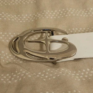 Vitt skärp med silverfärgat spänne - Snyggt vitt bälte med ett stort, ovalt silverfärgat spänne i metall. Bältet är stilrent och har flera hål för justerbar passform. Perfekt accessoar för att lyfta din outfit med en clean och modern look.
