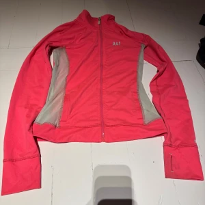 Rosa träningsjacka från Abercrombie - Sportig rosa träningsjacka från Abercrombie & Fitch med grå meshpaneler på sidorna. Jackan har hel dragkedja, hög krage och tumhål i ärmsluten. Perfekt för löprundan eller gymmet, med snygg logga på bröstet och ärmen.