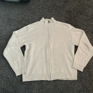 Beige kofta från Lindex med dragkedja - En stilren beige kofta från Lindex i storlek 146/152. Koftan har hel dragkedja framtill, ribbade muddar och hög krage. Perfekt för lager-på-lager och enkel att matcha med andra plagg. Materialet är mjukt och skönt, passar för både skola och chill. Strlk 152 men passar större!