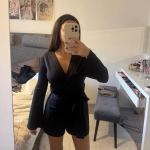 Byxdress  - Snygga svarta playsuit med brett knytband i midjan. Modellen är högmidjad och har en stilren, loose passform som ger en cool vibe. Materialet känns mjukt och har ett lätt fall. Perfekta för dig som gillar minimalistisk stil och vill ha något som sticker ut.