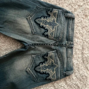 Blå jeans med broderade bakfickor - Snygga blå jeans som liknar Miss Me med unika broderade detaljer på bakfickorna och dekorativa sömmar längs benen. Jeansen har bootcut-modell och klassisk femficksdesign. Perfekta för dig som vill sticka ut med coola detaljer och bekväm passform. Skulle säga typ 90 cm innersida lår och sitter väldigt lågmidjat.(jag är 170) passar perfekt