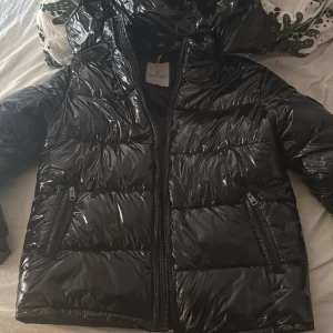 Svart pufferjacka från Moncler - Snygg svart pufferjacka från Moncler med glansig finish och klassisk logga på ärmen. Jackan har huva, två dragkedjeförsedda fickor och quiltad design. Perfekt för kalla dagar och riktigt street. Fodrad och med coolt serietryck på insidan.