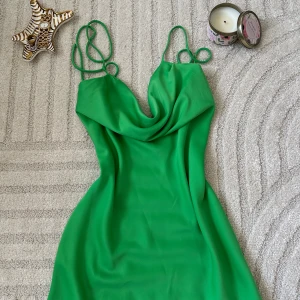 Grön kort klänning med tunna axelband med öppen rygg - Snygg grön kort klänning med tunna axelband och draperad urringning. Klänningen har en enkel, figurnära passform och är tillverkad i ett lätt, slätt material som faller fint. Perfekt för dig som vill sticka ut med en färgstark look.